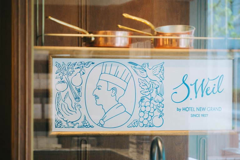 メインビジュアルにはサリー・ワイルさんの横顔に、ニューグランドを象徴するフェニックス、大階段のフルーツバスケットのオブジェが柔らかなタッチで描かれる。