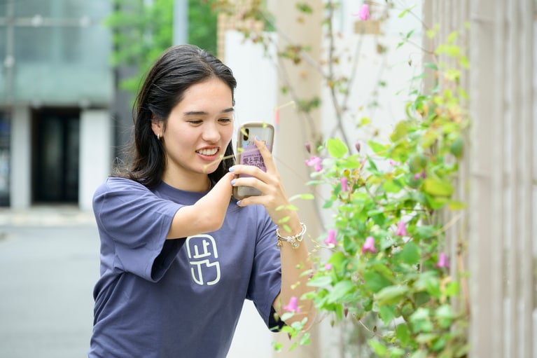 街中で見つけた花をスマートフォンで撮影する一ノ瀬メイさん。