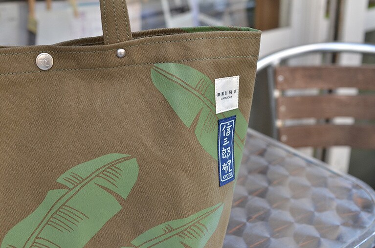 オリーブグリーンの帆布にバナナの葉の柄を染めた商品には『信三郎布包(しんざぶろうかばん)』のブランドネームと樂園百貨店、2つのダブルネームがつけられている。