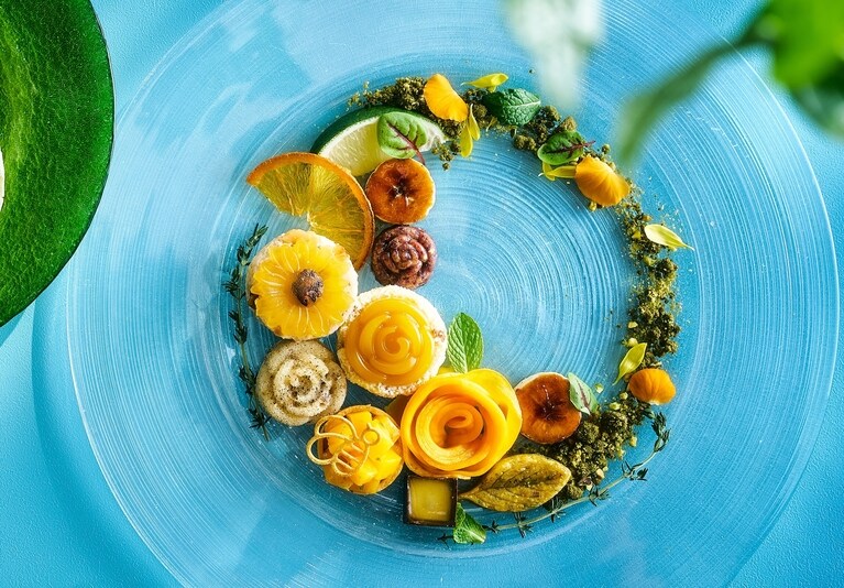 マンゴーとパイナップルにココナッツオイルなどを加えマンゴーのジュレで夏の花をデザインした「マンゴーとパイナップルのココナッツケーキ」などが美しくデコレーションされた、スイーツプレートのイメージ。