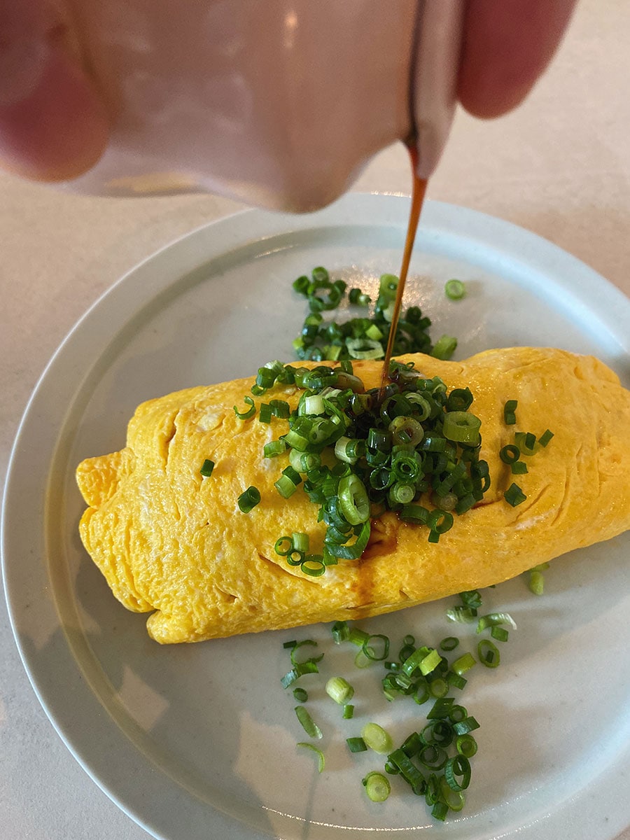 【焦がし醤油のケチャップライスが主役のオムライス】(7) 卵を1人分ずつ作る。ボウルに卵2個を割り入れたら菜箸でボウルの底をこするようにしてしっかり溶きほぐす。フライパンにバターを熱し、溶き卵を一気に流し入れて全体をサッと混ぜて半熟状になったらチキンライスの半量を中央にのせてフライパンの片側に寄せ、皿に返して盛りつける。小ネギと醤油をトッピングして完成。