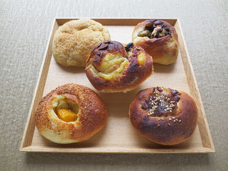 左上から時計回り：「メロンパン」275円、「オーガニックバナナとチョコチップ」275円、「あっさり粒あんぱん」275円、「フレッシュマンゴーと豆乳クリーム」275円。中央は、オリジナルのチャイスパイスが香る「豆乳クリームパン・チャイ」275円。