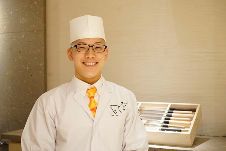 店主の村井成至さん。写真を撮るためにマスクを外していただいたら、現れたのはとびきり爽やかな笑顔！　胸の刺しゅうにも使われている店名のロゴは、父・隆さんが、「天ぷら 近藤」の油と墨を混ぜて書いたという。