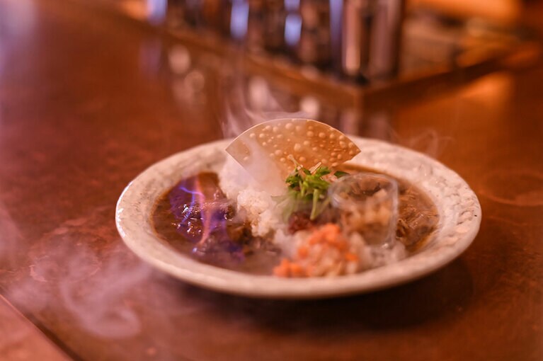 インドカレーの「ポークビンダルー」をベースにアレンジし、酸味を効かせた『メラメラ』と、カツオだしの旨みが豊かで少しスモーキーな『モクモク』のあいがけ1,500円。『メラメラ』は、カレーの表面から炎があがった状態で運ばれ、『モクモク』には燻煙が吹き込まれた食用シャボン玉が添えられる。途中で2種類のカレーを混ぜ合わせれば、また味わいが変わり、3つの味が楽しめる。