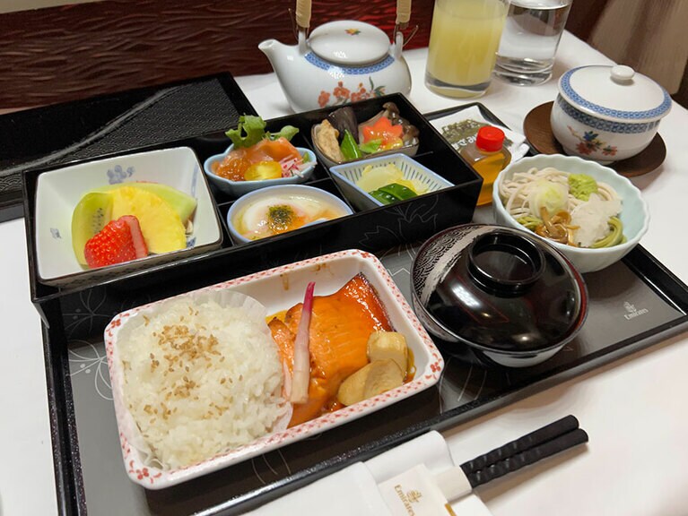 朝食の懐石弁当。揚げ出し豆腐、ナス、しめじ、サーモンのマリネのキュウリと大根添え、サーモントラウト照り焼き、エリンギとお漬物、味噌汁、大根となめこが乗った蕎麦と抹茶うどん、フルーツ。