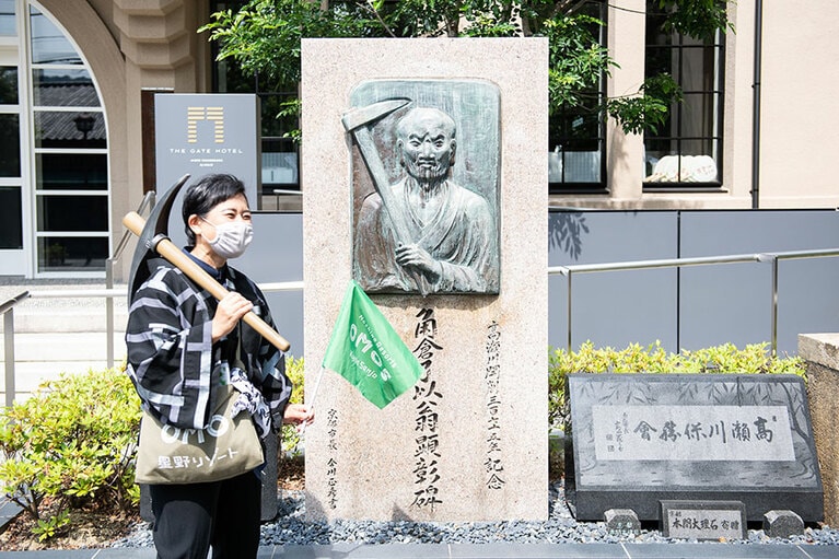 角倉了以の胸像前で、小道具を片手に話す八十田さん。高瀬川の開削はツルハシなどを使い人力で行われた。