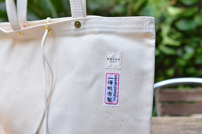 生成の無地商品には『一澤帆布製』のネーム。カバンは良質の天然帆布を使い、昔と変わらず丁寧な手作業ですべて製造されている。シンプルで使い勝手が良い頑丈なかばんは、メイドインジャパンのハイクオリティとして国内外に多くのファンを持つ。