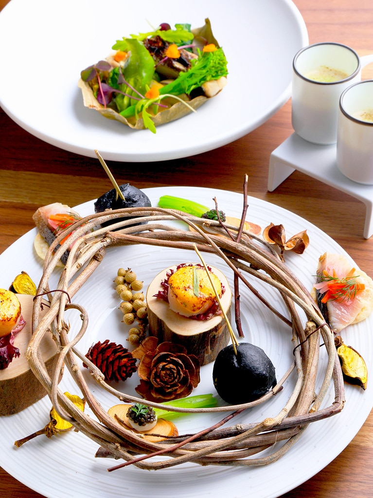 手前のアミューズはホタテのオランデーズソース焼き、リンゴ酢でしめた八戸産〆サバ、フォアグラと奈良漬入り竹炭ボウル、ミニかぶのキャビアのせ、かぶのポタージュ(写真は2人前)。奥のメインの魚は島根県産のタラ。柿の葉で包み、豊澤酒造の日本酒で蒸しあげる。※いずれもディナー 10,000円より。