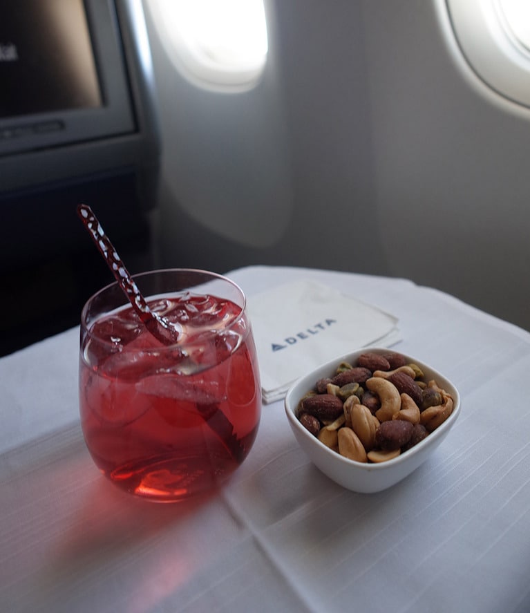 ジンをベースに、クランベリーアップルジュース、ジンジャーエールを加えたデルタ航空特製サンライズ・カクテルと温かいナッツ。カクテルは季節ごとに種類が変わる。このマドラー、プラスチック製のものから環境に配慮した竹製と樺の木製マドラーへと順次交換されている。 