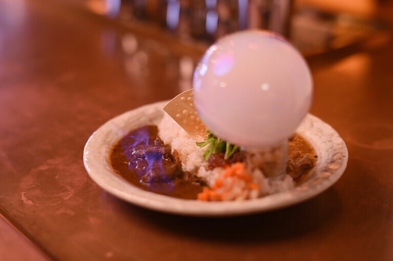 インドカレーの「ポークビンダルー」をベースにアレンジし、酸味を効かせた『メラメラ』と、カツオだしの旨みが豊かで少しスモーキーな『モクモク』のあいがけ1,500円。『メラメラ』は、カレーの表面から炎があがった状態で運ばれ、『モクモク』には燻煙が吹き込まれた食用シャボン玉が添えられる。途中で2種類のカレーを混ぜ合わせれば、また味わいが変わり、3つの味が楽しめる。