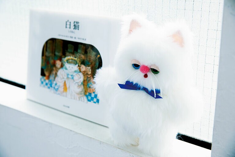 プレゼントは、2人ともがファンのpienilusikkaさんにオーダーしたぬいぐるみ「もけもけ」。オモムロニ。さんの愛猫「アイビー」の真っ白な毛とオッドアイに、やはり大好きな横浜DeNAベイスターズのイメージカラーリボンを巻いてほしいと注文。「もけもけ」 9,000円(参考価格)／pienilusikka　※現在はメールでのオーダーは休止中で、イベントなどでのみ受付。奥はおまけプレゼントの絵本『白猫』(ほるぷ出版)。