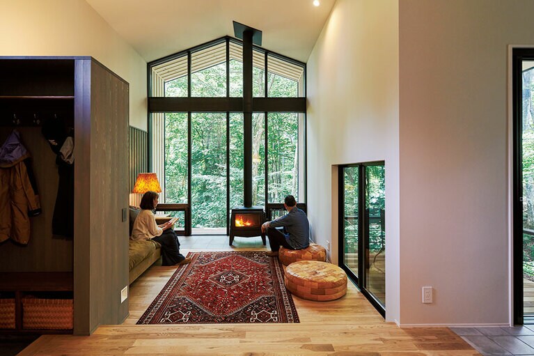 目の前の木立の眺めを最大限に取り込む、リビングの大きな窓。反対に写真右手のテラスにつながる窓はやや頭を下げて出入りするくらいの小ささだ。「窓が大きすぎたり、多すぎると落ち着かないんですね。これくらいがちょうどいい」(Kさん)。薪ストーブの炎と間接照明の灯りが、さらにくつろぎ感をもたらす。