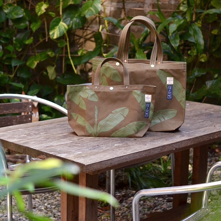 バナナの葉の柄は、沖縄出身のアーティスト「MIREI」氏によるデザイン。樂園百貨店に併設する樂園カフェでもメインビジュアルとして店内に描かれているバナナの木は、沖縄ではポピュラーな植物だ。バナナの葉には、ゆったりとした沖縄の風を感じさせ、「お店や商品を包む」といったメッセージを込めている。