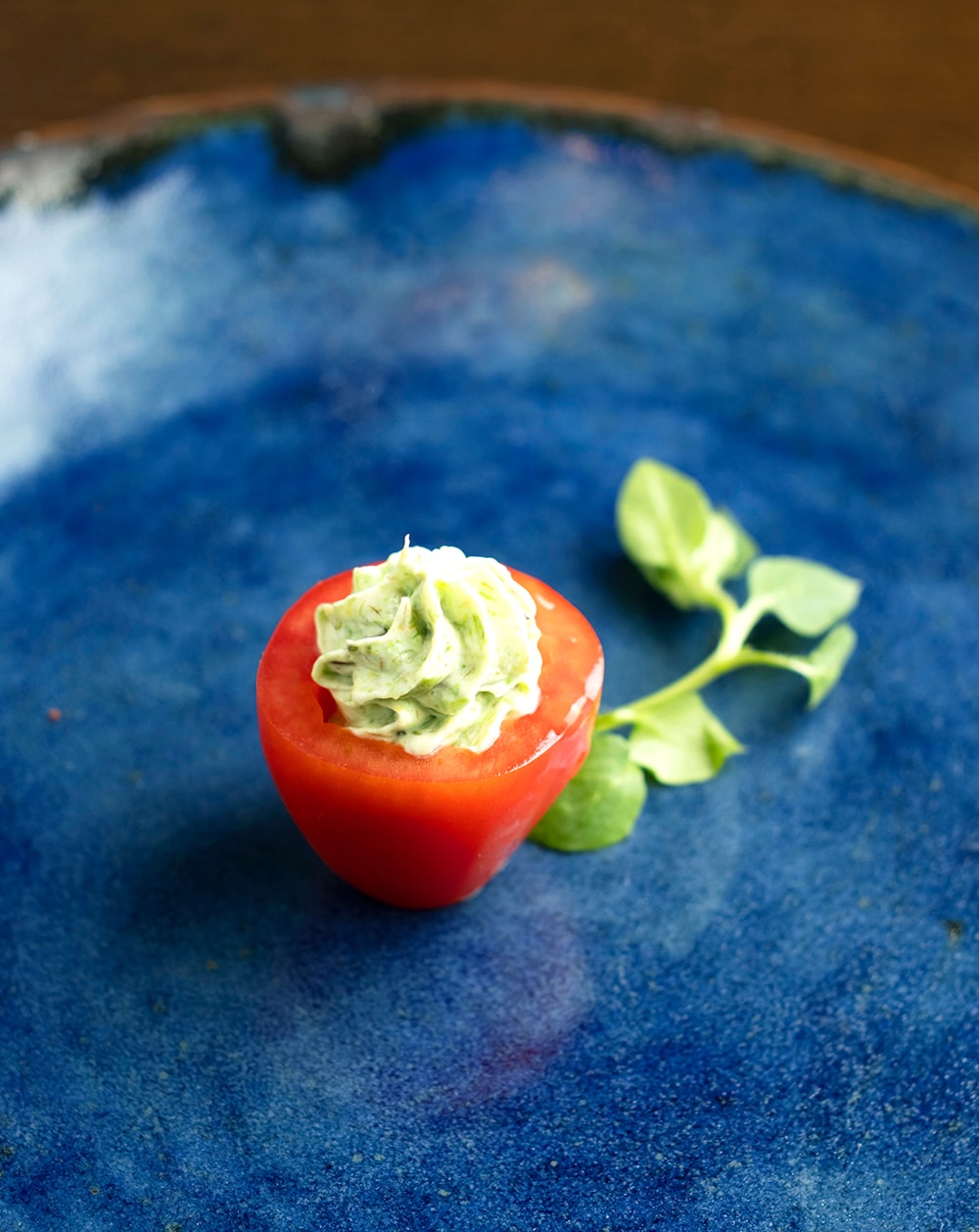 フキノトウを詰めた真っ赤なミニトマトをあえて鮮やかなブルーのお皿に。器は竹田市内の「無風窯」高木逸夫さんの作品。