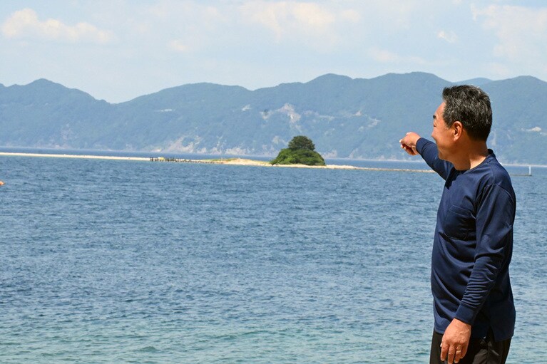 色浜の港から水島を指さす古川さん。