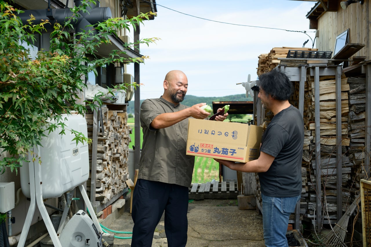 採れたてのナスを詰めた段ボールを手に、「佐藤大農園」の佐藤さんがレストランへお届け。井上シェフはその艶を見つめ、とっておきのひと皿を思い描きます。