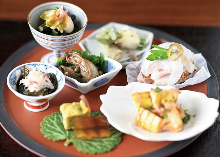 前菜盛り合わせ。胡麻の餡をかけたわらびとセリのお浸し、ウドに真鯛の昆布締めを巻いた天ぷら、自家製のハムをのせたブルスケッタ、ホンズワイガニの解した身をのせている根もずく、牡蠣の有馬煮など、旬を詰め込んだ7つの小皿が並びます。