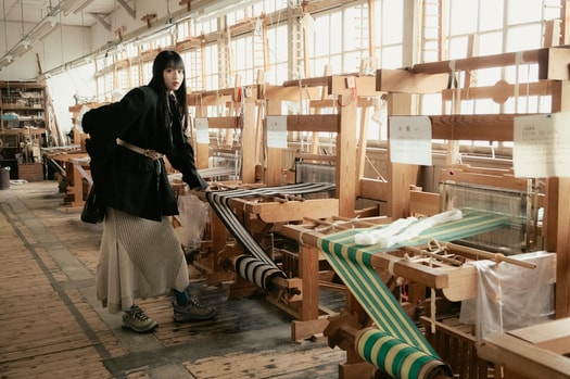 建物内では秩父銘仙の歴史や展示品を見ることができるほか、型染めや織りの体験も可能。木製の織り機や設備はすべて現役で使われているので、職人が織りを行う姿に出会えることも。菊池さん着用：ターギー フォー ウォータープルーフ ¥24,200／キーン（キーン・ジャパン）、ジャケット 96,800円、ベルト 36,300円／共にフミエタナカ（ドール）、ニットトップス 57,200円、ニットスカート 52,800円／共にオープン セサミ クラブ バッグ 37,400円／ルメメ、タイツ／スタイリスト私物