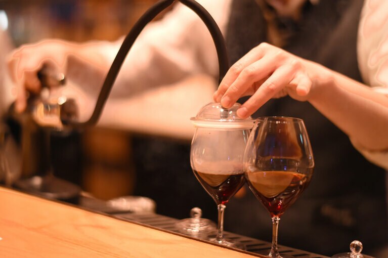 ウイスキー「シーバスリーガルミズナラウッドリザーブ」にベルモット、国産カシスリキュール、黒蜜を合わせて、スモークガンで燻製の香りづけ。それを自ら手元で温かいアメリカーノに注ぎ込むことで完成する。焚火の香りを連想させる、冬季限定のホットカクテル。