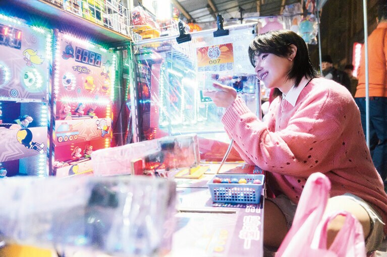 【台北】寧夏夜市（ニンシャーイエシー）全長300メートルほどの道に屋台が並ぶ地元民に愛される夜市で、童心に返って、おもちゃのピストルで的当てゲーム。「子どもの頃、家族と一緒に来た思い出の場所です。人混みが苦手で、当時はこんなに楽しめなかった！」ニット、中に着たシャツ、ハーフパンツ、ネクタイ／ジバンシィ（ジバンシィ ジャパン）　ピアス／スタイリスト私物