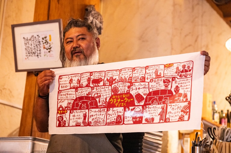 お店オリジナルの「ウチナーウヮー（沖縄の豚）すごろく」手ぬぐいを使いながら豚食文化を説明する満名さん。店長の人柄もお店の魅力の一つだ。