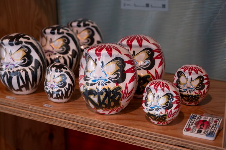 達磨は、まず叶えたい願いを込めて左目を、願いが叶った際には右目を入れる。「満願」したらさらなる飛躍を祈る魔除けとなってくれる。オリジナル達磨は赤と青の2色。万博限定品。