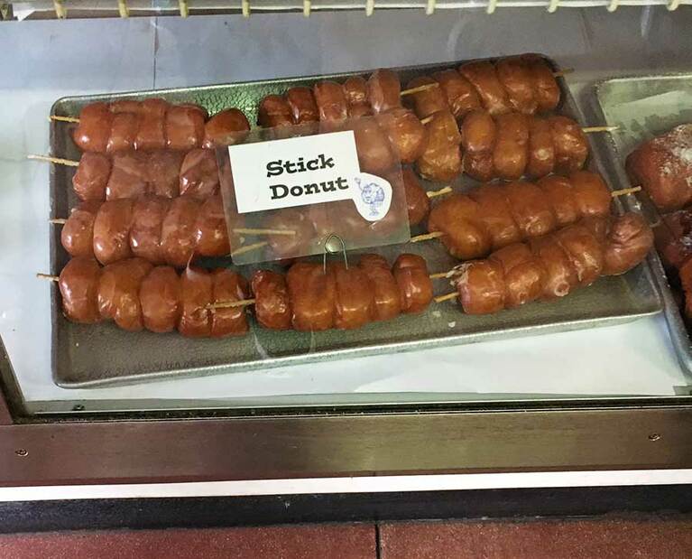 コモダベーカリーの名物「スティックドーナツ」。砂糖がけ＆串刺し！　素朴なんだかワイルドなんだか。これがいちばん有名なんだけど、なぜか店のマスコットキャラクターはクリームパフ(ロゴの右下にいる)。もちろんクリームパフもおいしいよ！