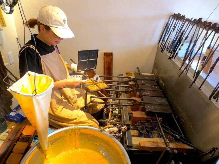 入り口すぐの焼き場で、おせんべいを手焼き。生地は、カステラと同じ小麦粉と卵、砂糖。昔からの職人は、床にあぐらをかいて焼き型を回すが、絵三子さんは椅子に座って作業し、腰の負担を軽減。女性でもできるスタイルに。